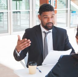 Businessman gesturing and talking with partner in outdoor cafe. Business man sitting and talking with woman who is sitting back to camera with building glass wall in background. Negotiations concept.