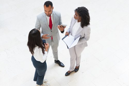 Diverse business team meeting in office hallway. Business man and women standing in circle, holding tablet and documents and talking. Teamwork concept